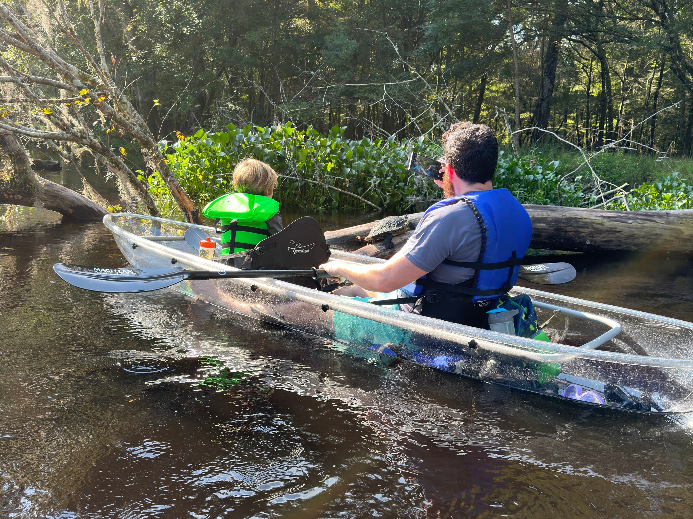 Two people canoeing in a clear boat on a river, surrounded by trees.
