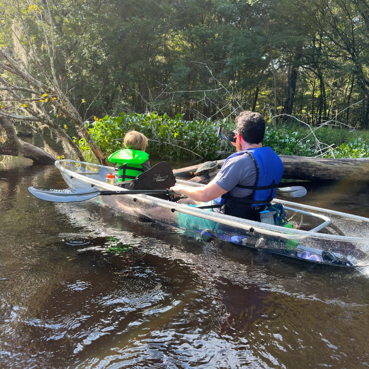 Two people canoeing in a clear boat on a river, surrounded by trees.