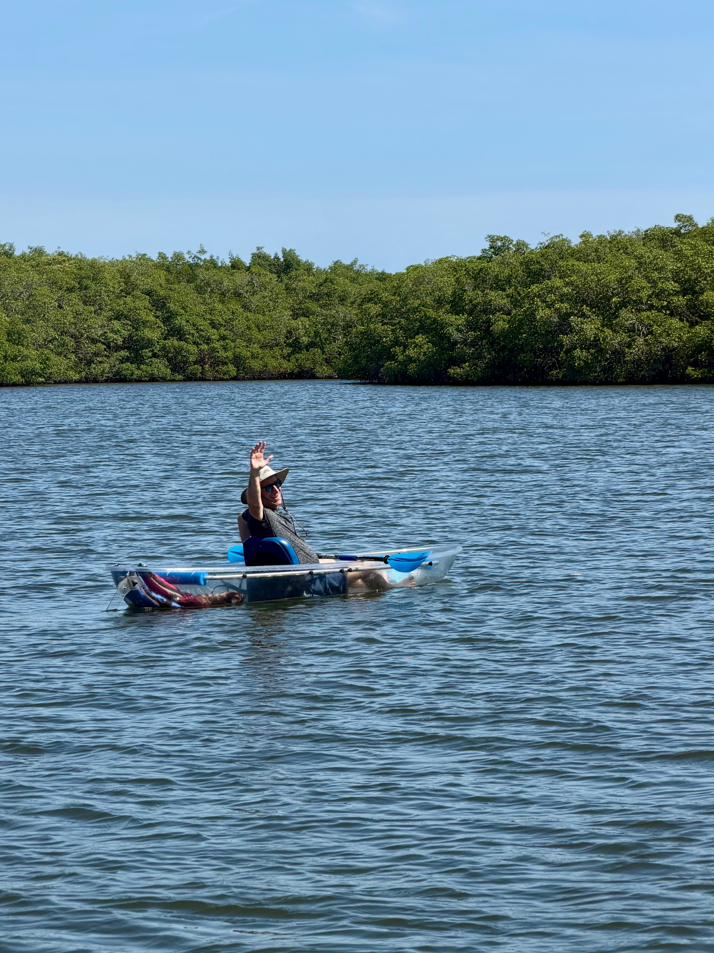 Person in a hat waves while kayaking on a calm river surrounded by dense greenery.