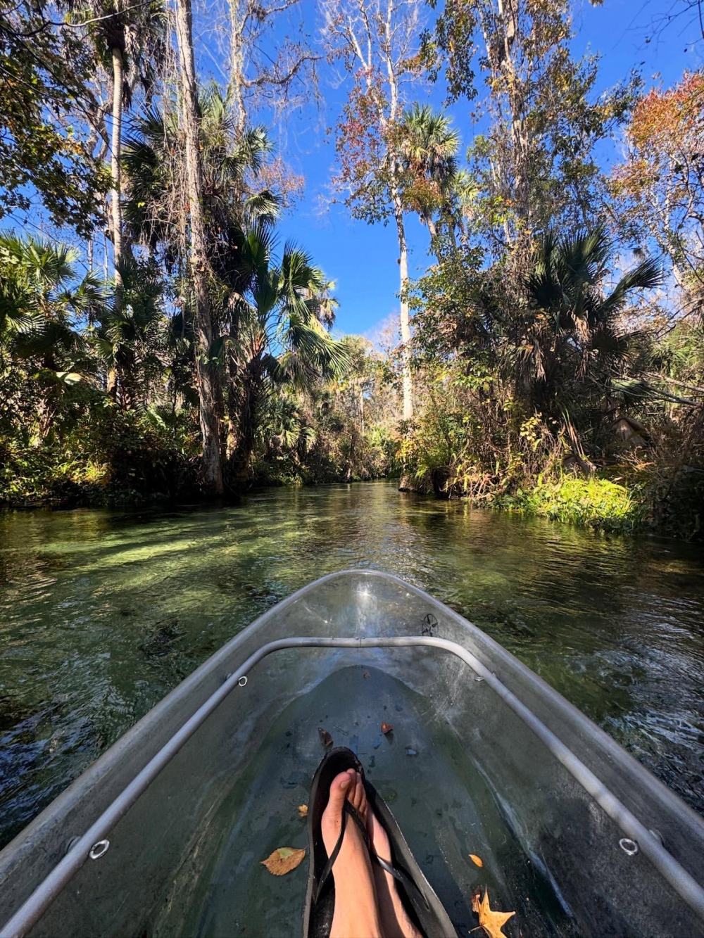 Person's feet in a hammock kayak on a calm, forested river under a clear blue sky.
