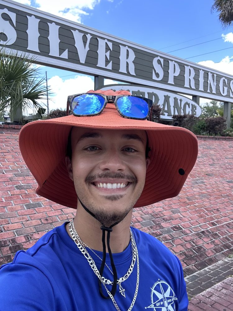 Person in orange hat smiling, standing by a sign that reads 'Silver Springs Entrance'.