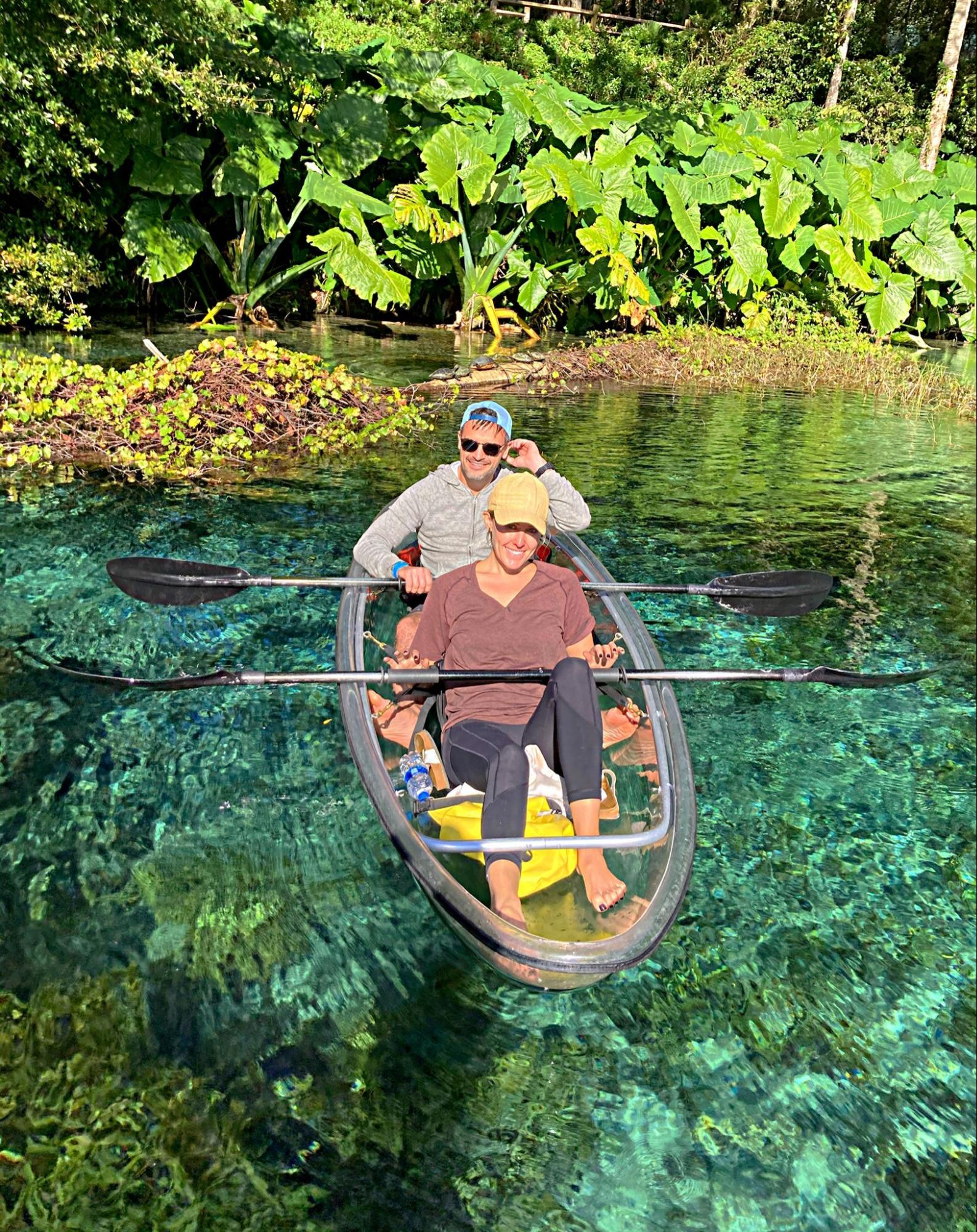 Two people smiling in a clear kayak on a turquoise river surrounded by lush greenery.