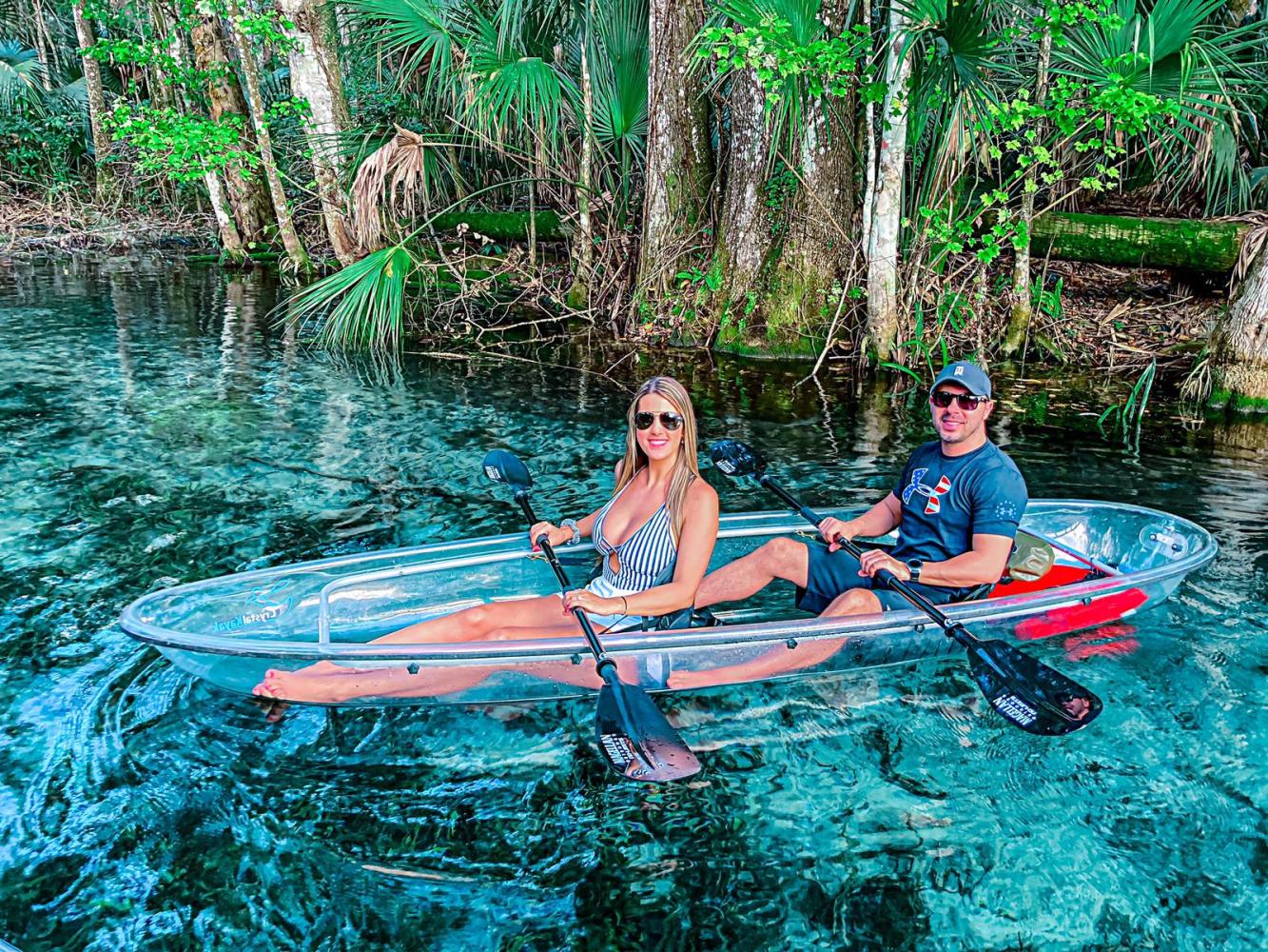Two people kayaking on clear water surrounded by trees in a glass-bottom kayak.