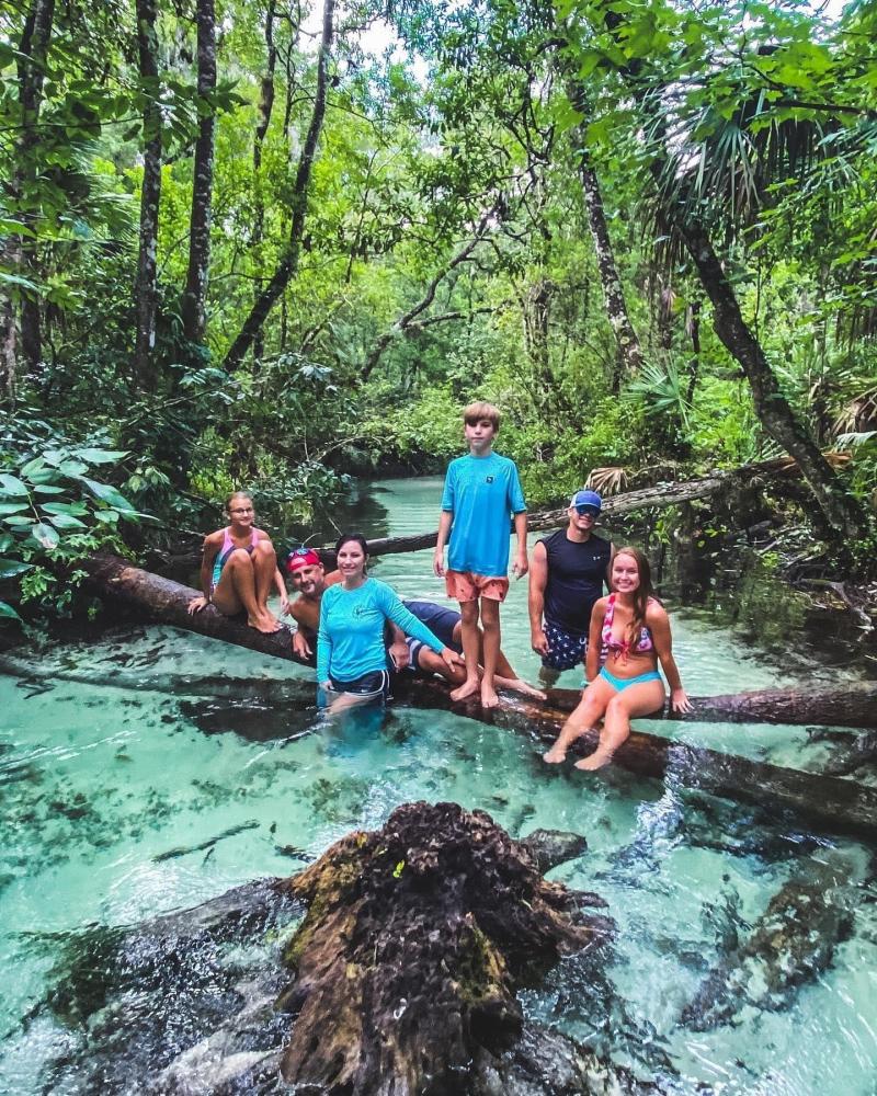 Group of people relaxing on logs in a clear forest stream.