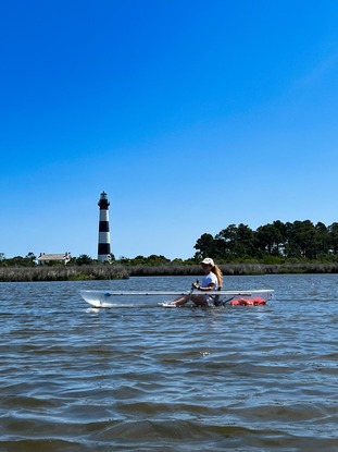 Person kayaking on water near a lighthouse under a clear blue sky.
