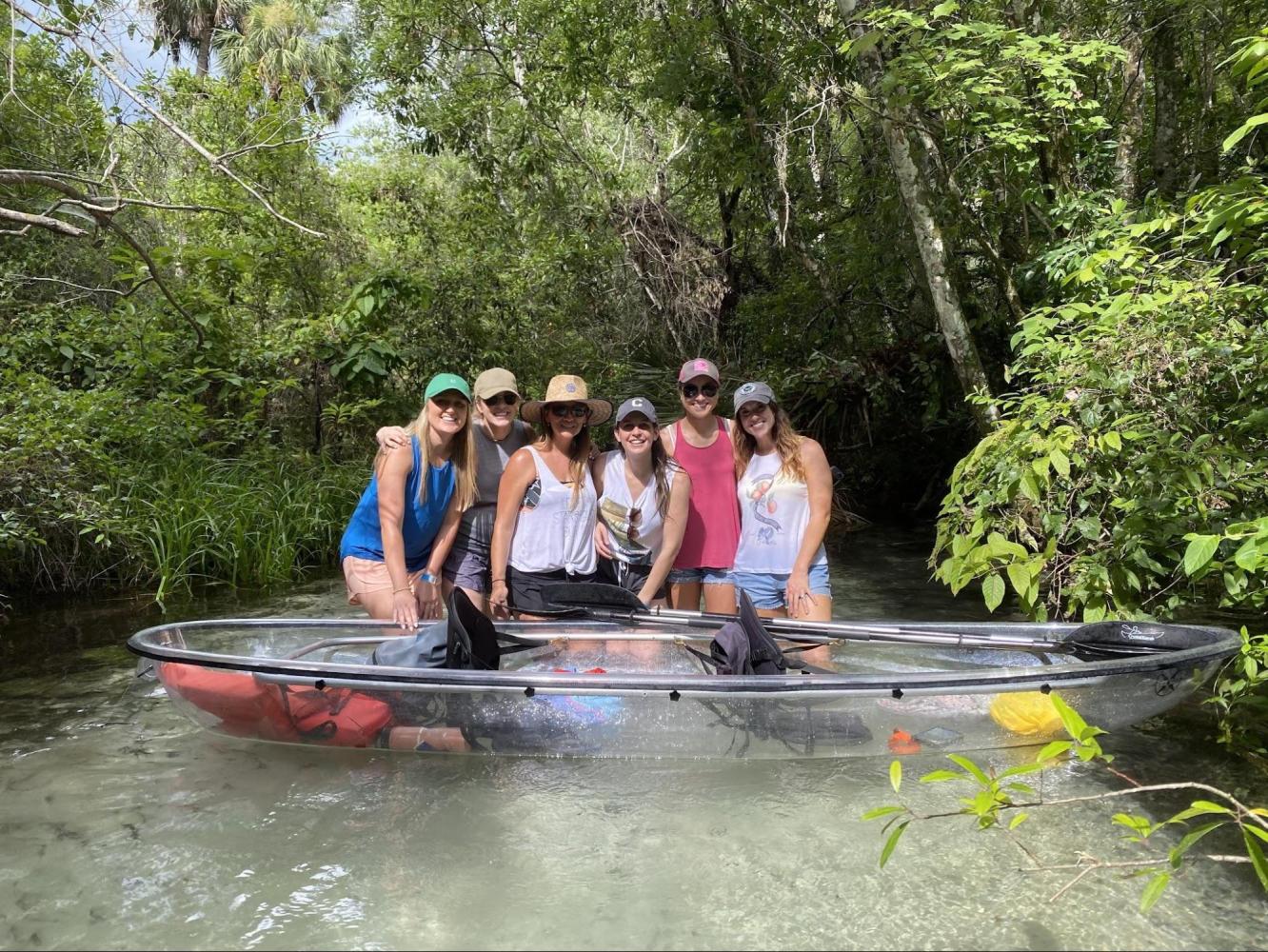 Six people pose in a see-through kayak on a lush, narrow waterway.