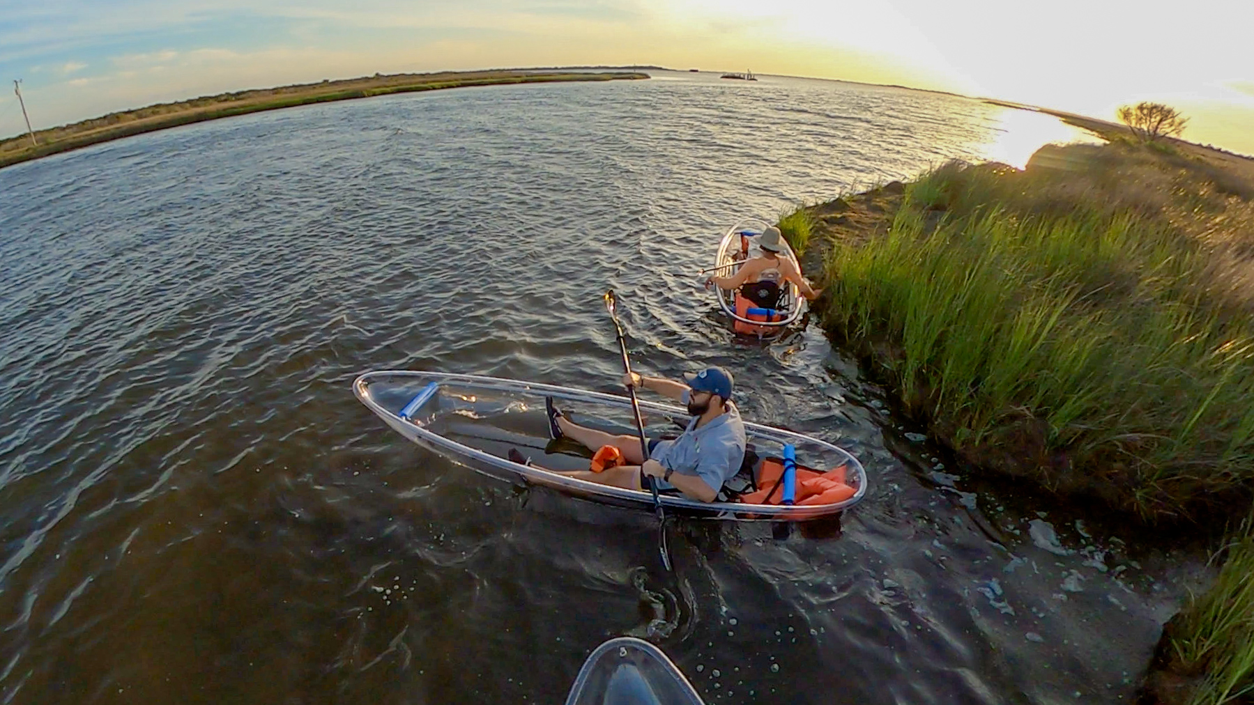 Two people in transparent kayaks on a river at sunset.