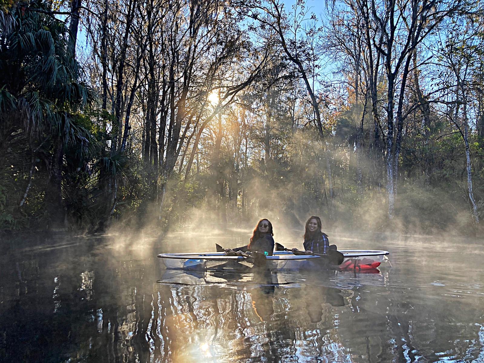 Two people kayaking on a misty river surrounded by trees at sunrise.
