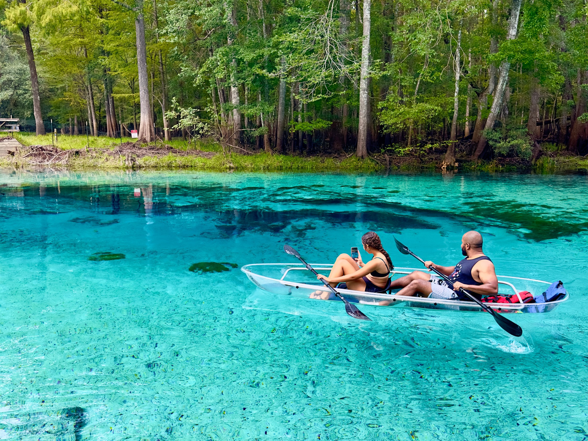 Two people in a clear kayak on a calm, blue river surrounded by trees.