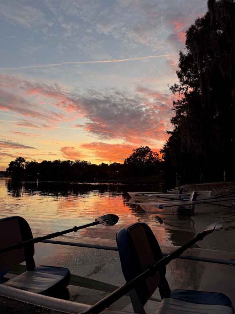 Sunset over lake with boats and trees silhouetted against colorful sky.