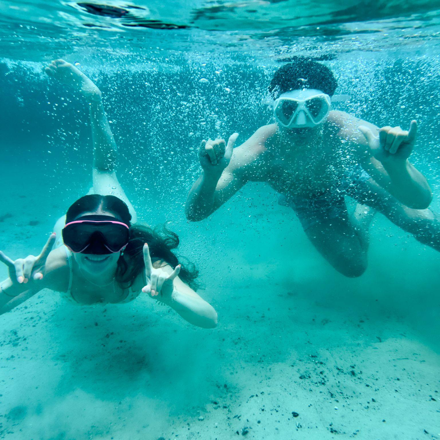 Two people underwater wearing goggles, making hand signs, and smiling.