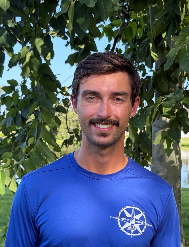 Man in blue shirt with compass logo, standing outdoors with trees in background.