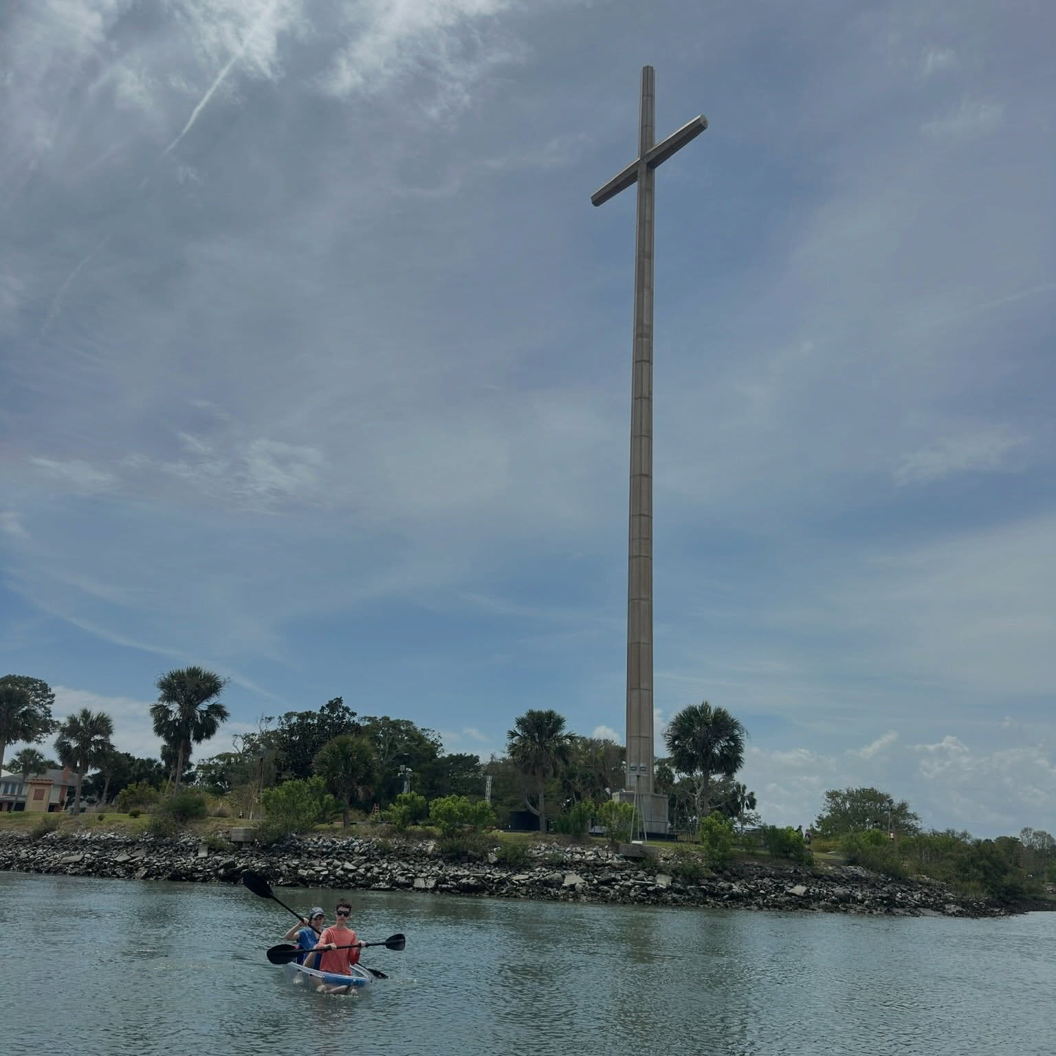 Two people kayaking on a river, large cross monument on the bank.