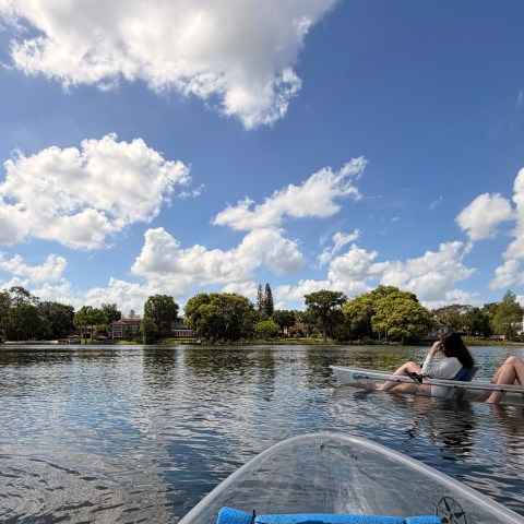 People in clear kayaks on a lake under a blue sky with clouds.