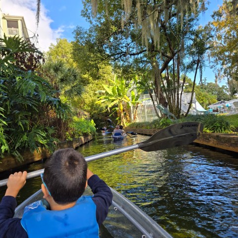 Child in blue life jacket paddling a clear kayak on a tree-lined canal.