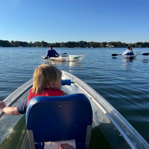Child in a kayak on a lake, accompanied by two adults in separate kayaks.