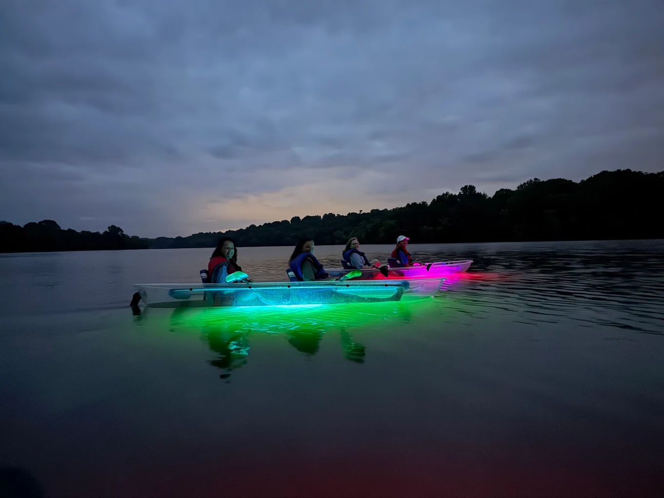 Kayakers on a lake with glowing green and pink lights at dusk.