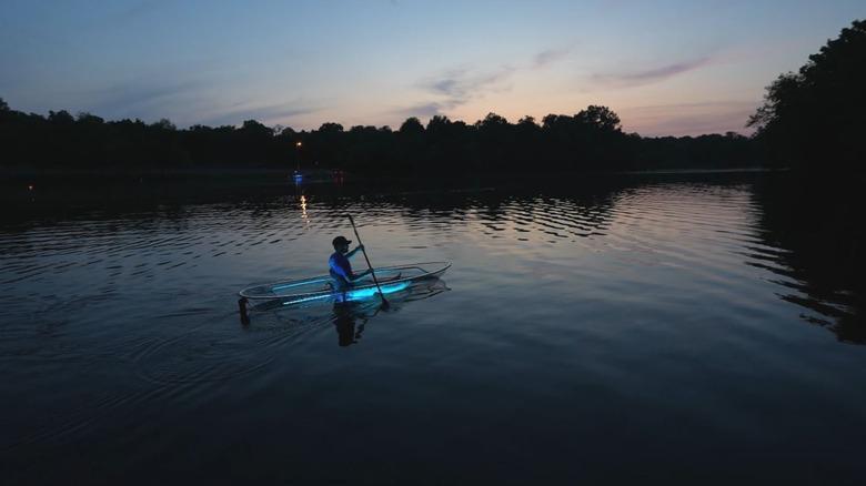 Person kayaking in clear boat on calm lake at dusk with glowing lights.