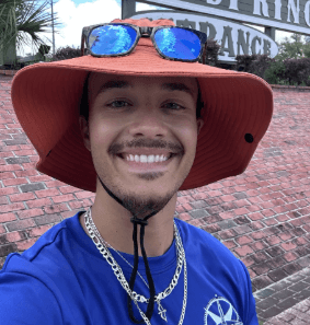 Smiling person with orange hat and sunglasses on top, wearing blue shirt and necklaces.