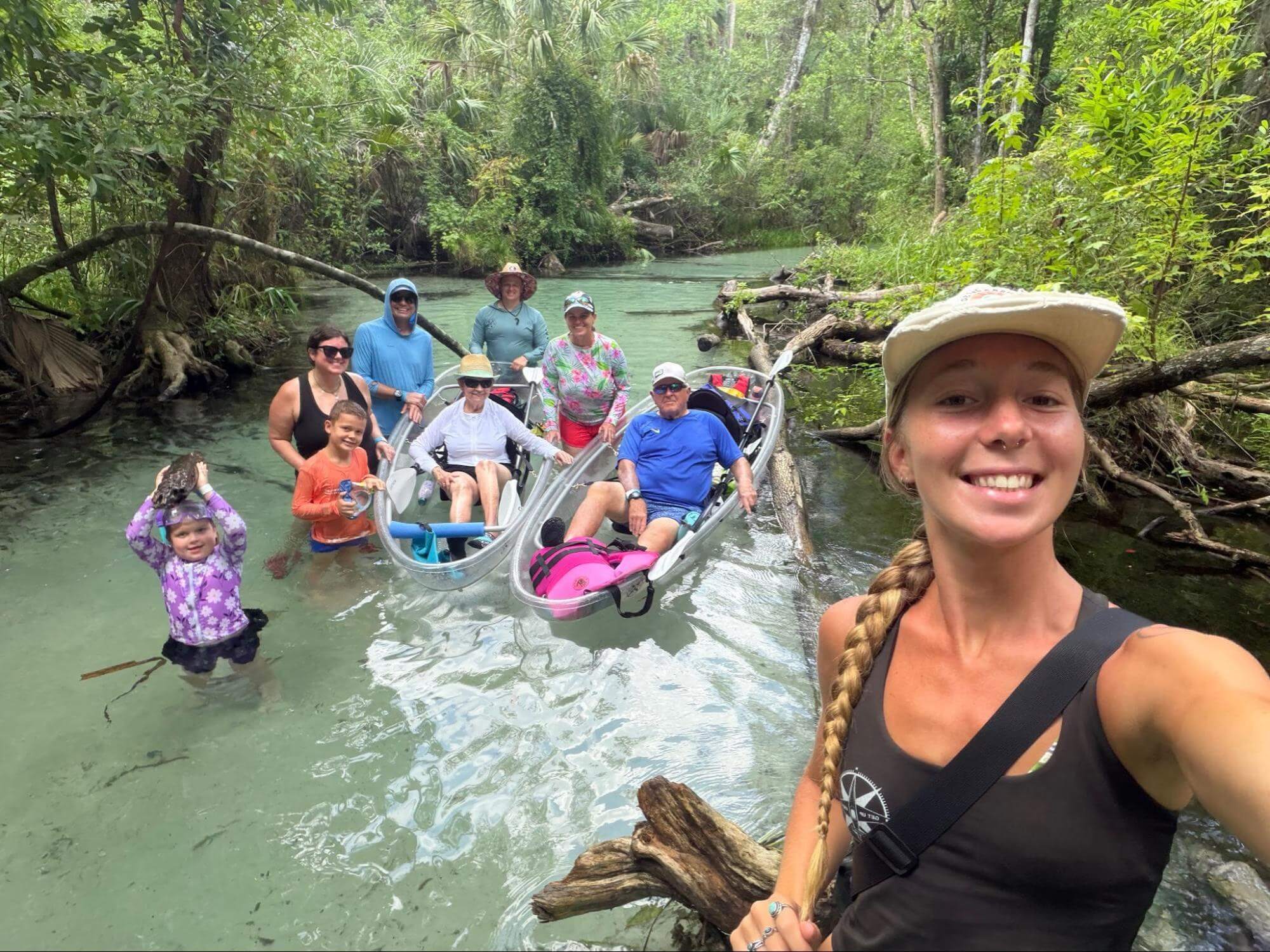 Group of people kayaking and standing in a shallow river in a forest.