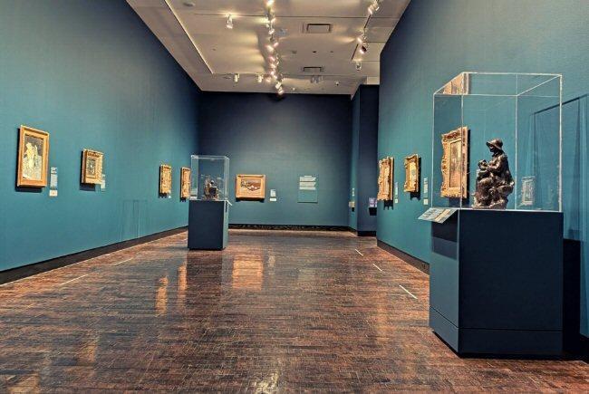 Art gallery with paintings on blue walls and sculptures in glass cases on wooden floor.
