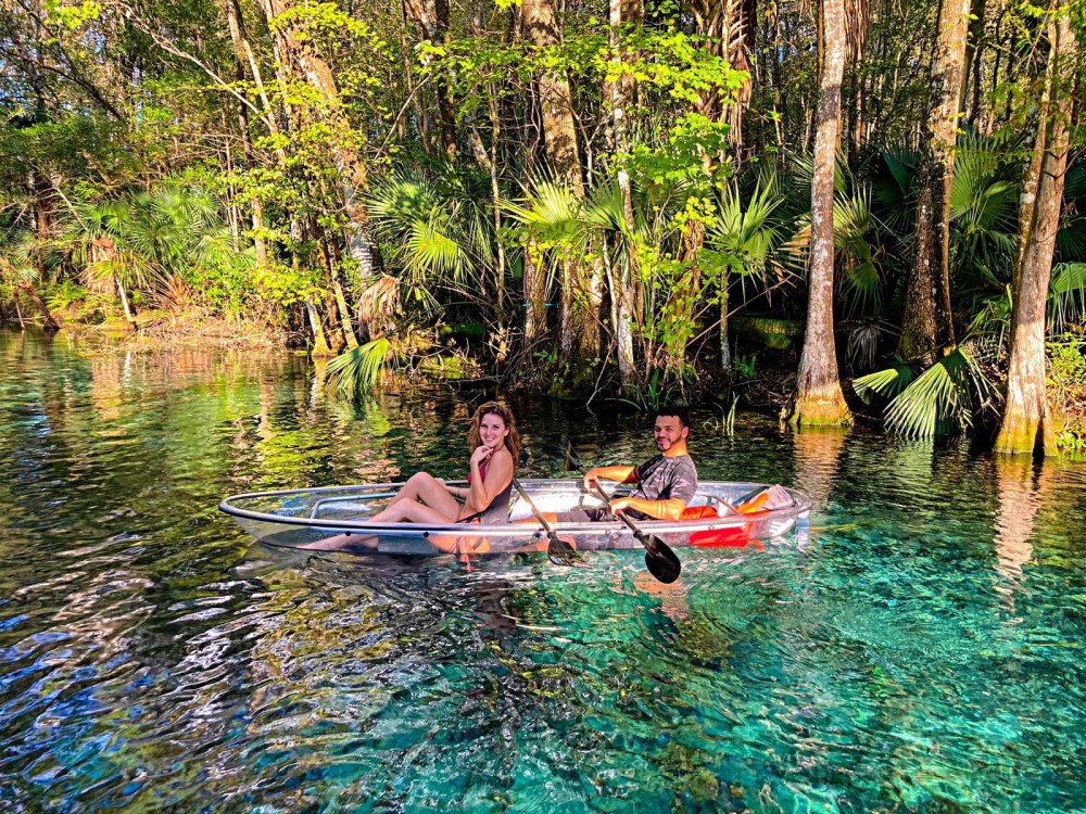 Two people in a clear kayak on a forested, clear blue water body.