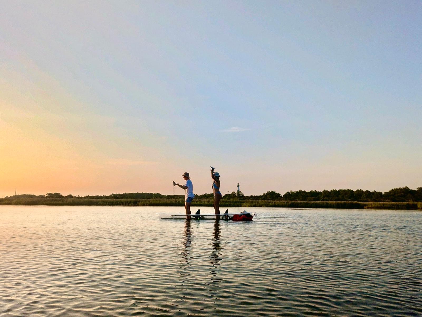 Two people paddleboarding on calm water at sunset, silhouetted against the sky.