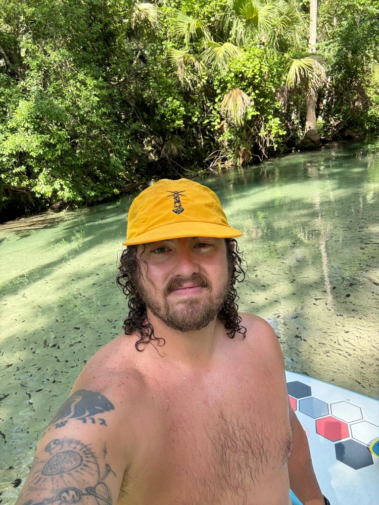 Person in a yellow cap kayaking on a clear, green river surrounded by lush trees.