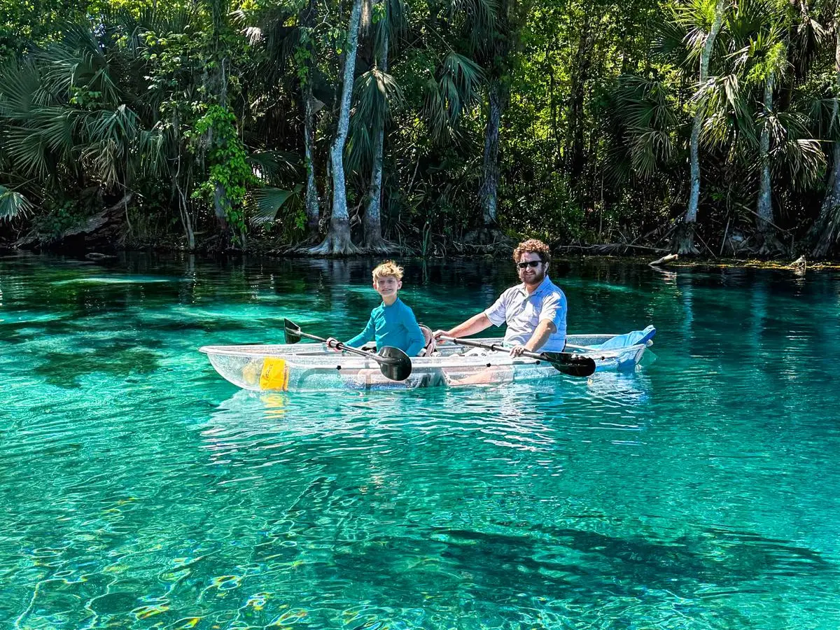 Two people kayaking on clear turquoise water surrounded by lush trees.