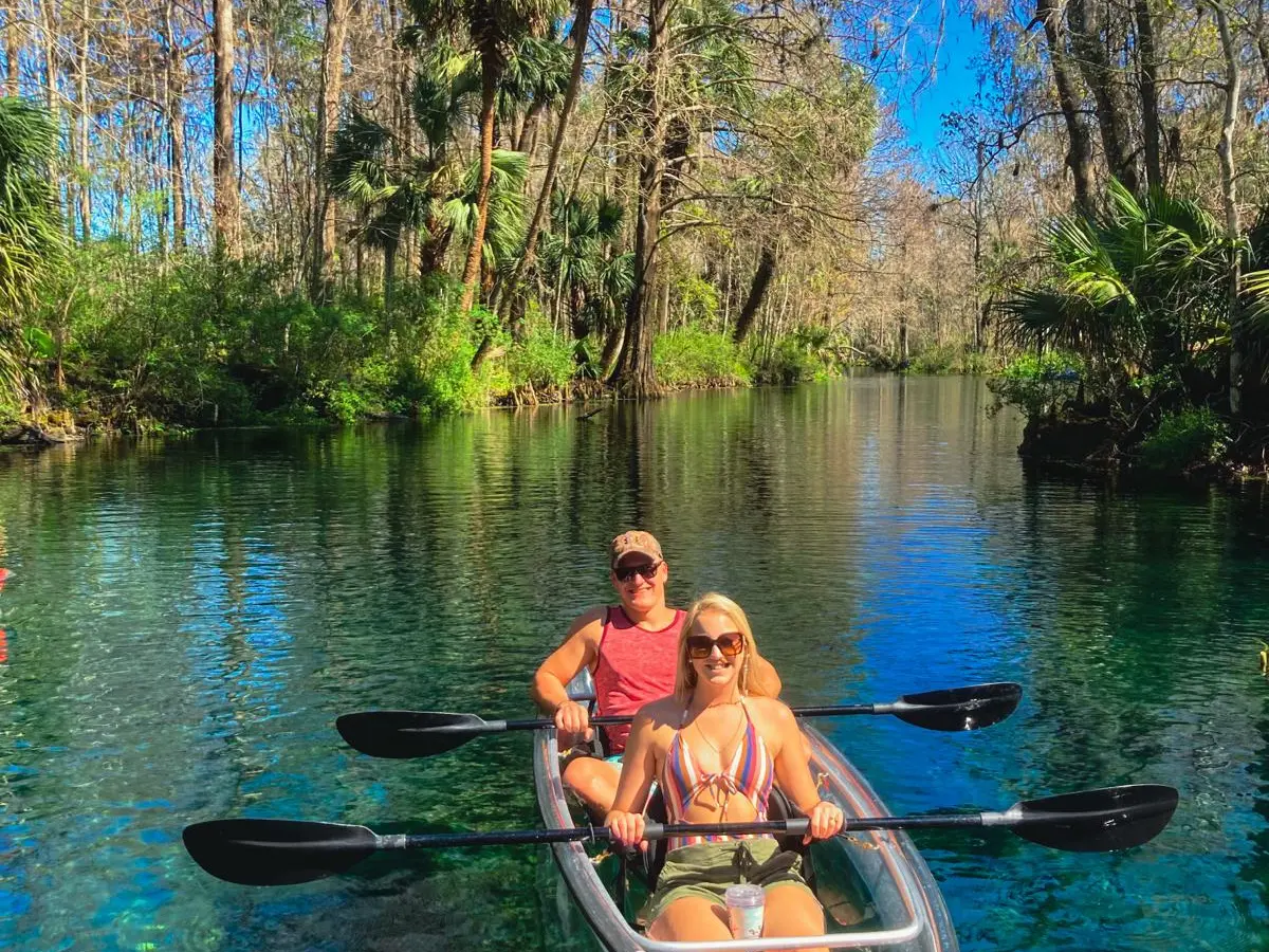 Two people kayaking on clear water in a lush, forested area.