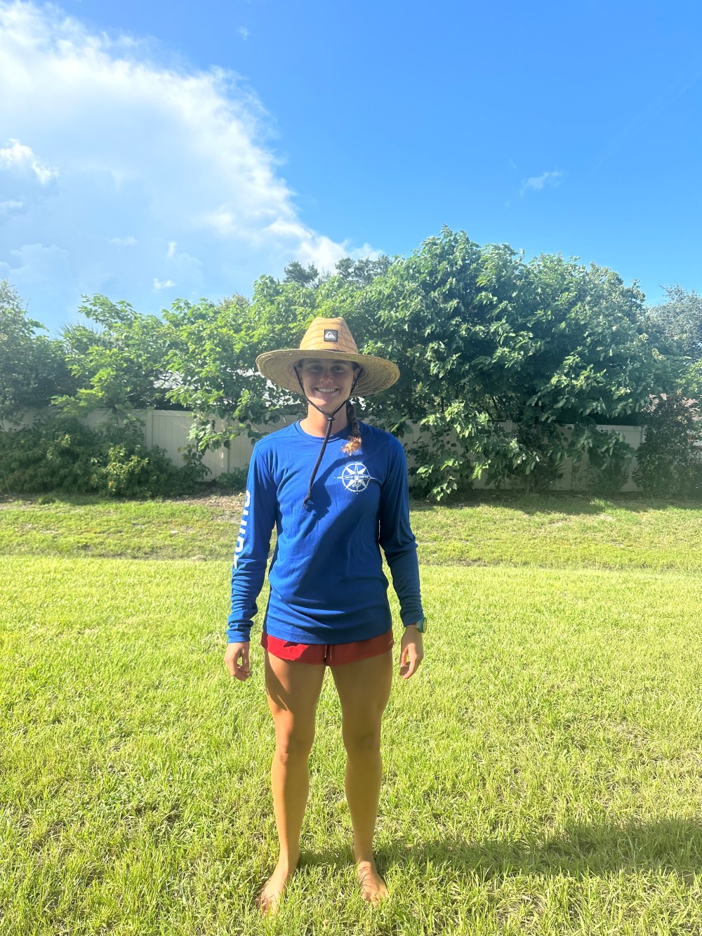Person in a blue shirt and straw hat standing on grass with trees and blue sky in the background.