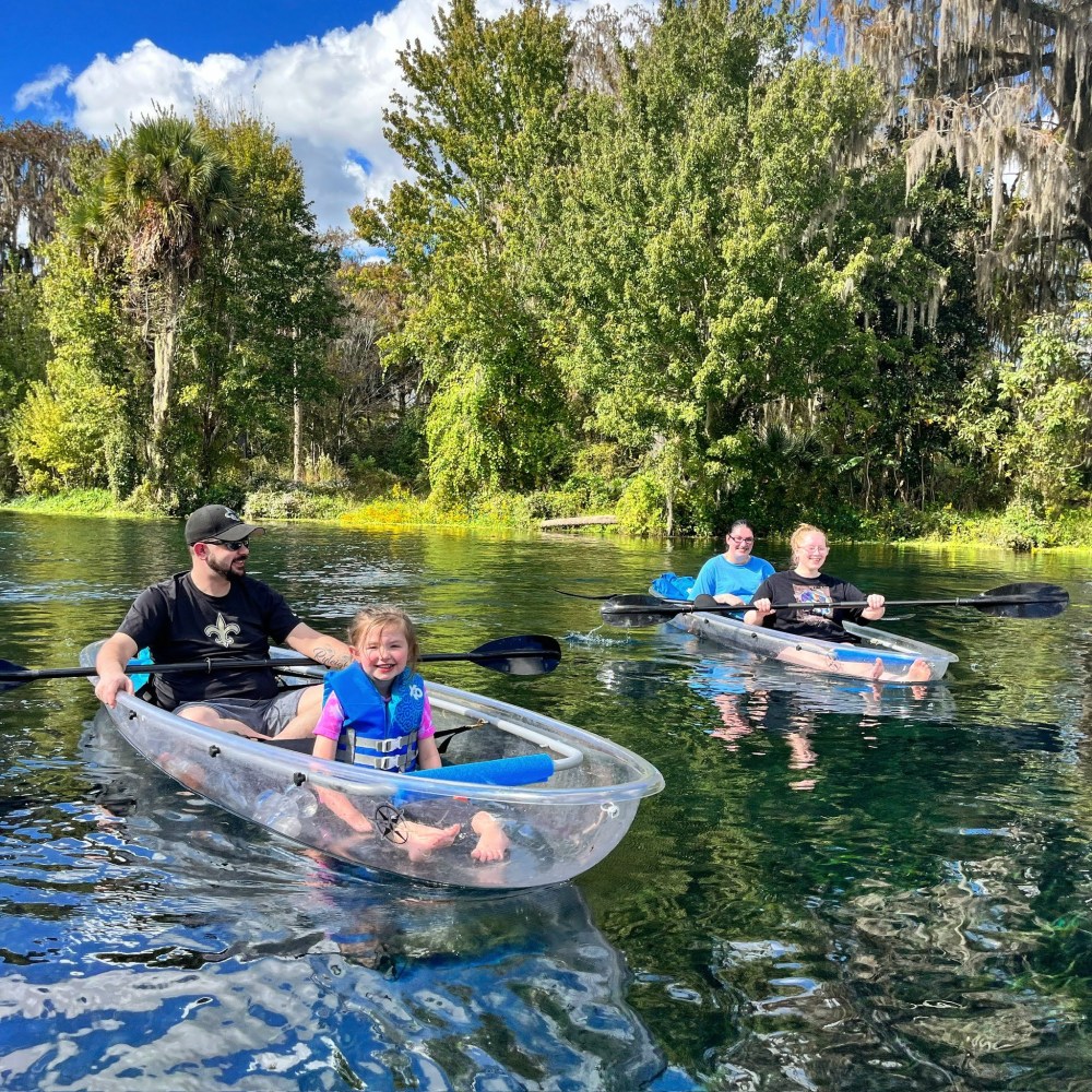 People kayaking in clear kayaks on a sunny river with trees in the background.