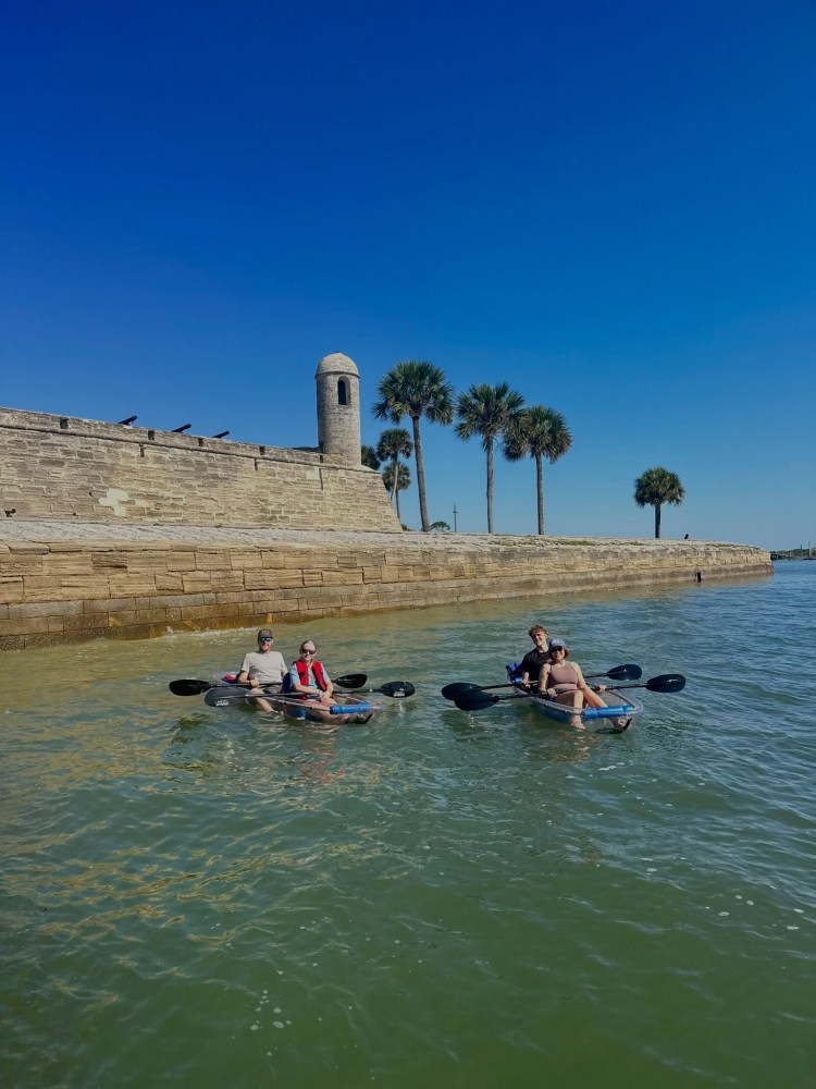 Three people kayaking near a stone fort with palm trees and a clear blue sky.