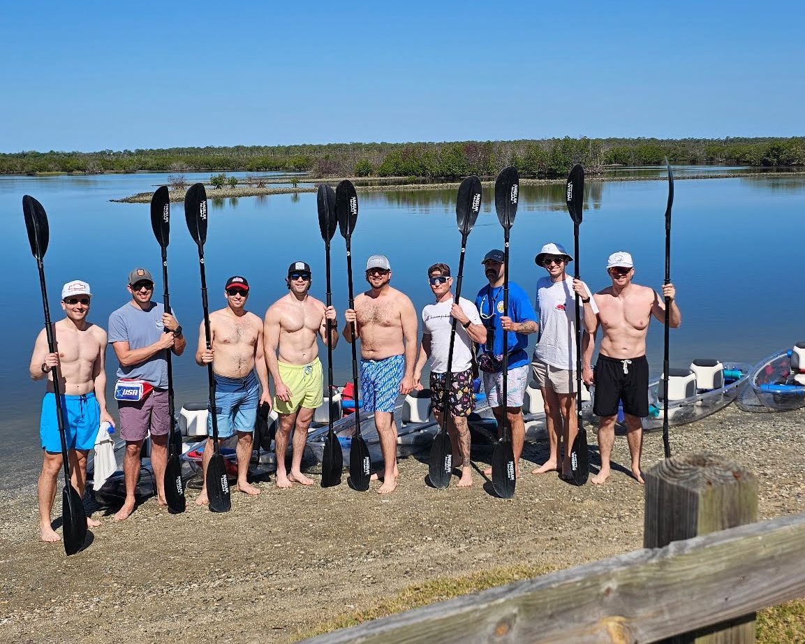A group of men holding paddles by a calm lake under a clear blue sky.