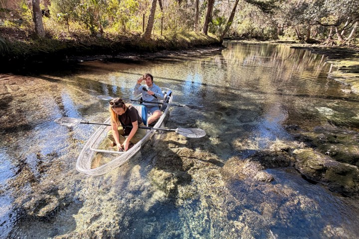 Two people in a clear kayak on a tranquil, wooded river.