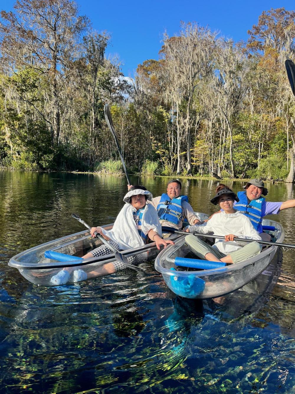 Four people in kayaks on a clear lake with trees in the background.